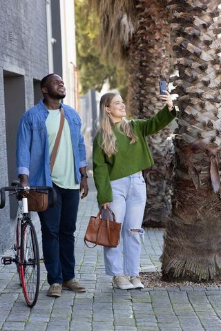 Couple strolling palm-lined urban walkway taking selfie while man holding bicycle