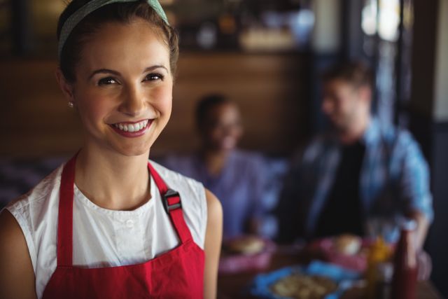Young waitress wearing red apron smiling at camera in a restaurant. Ideal for use in articles or advertisements related to hospitality, customer service, restaurant industry, and dining experiences. Can be used to depict friendly service, professionalism, and positive dining atmosphere.