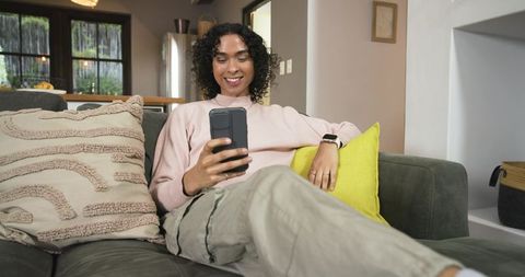 Nonbinary person relaxing on gray sofa using smartphone and smartwatch in cozy living room