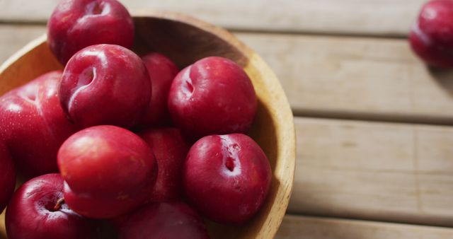 This stock photo depicts fresh, ripe red plums generously filling a rustic wooden bowl placed on a wooden surface. Ideal for use in food blogs, health and nutrition articles, fruit farming websites, and healthy lifestyle brochures to represent freshness and natural produce.
