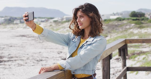 Young woman with curly hair taking a selfie on a wooden railing outdoors. She is wearing a denim jacket and standing against a backdrop of natural scenery with sand dunes and greenery. Ideal for use in social media, lifestyle blogs, travel promotions, influencer marketing, and capturing casual moments.