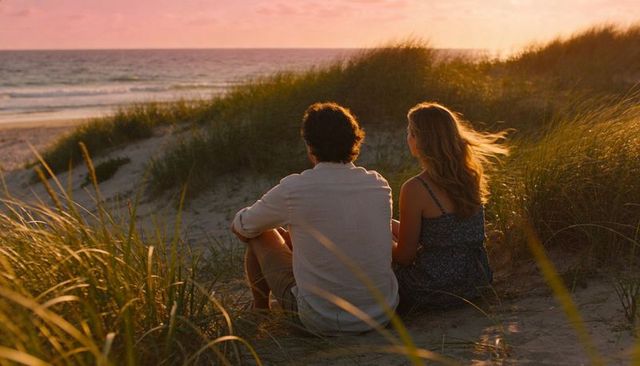 Couple sitting on sandy dune watching sunset ocean through sunlit beach grasses