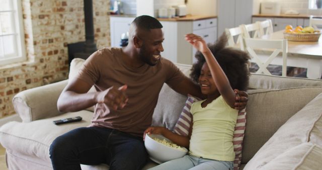 Father and daughter share a joyful moment watching a movie together at home. They are seated on a comfortable sofa with a bowl of popcorn. This image captures family bonding, casual living, and quality time spent together. Ideal for use in advertisements, articles, and promotions related to family life, parenting, home entertainment, and leisure activities.