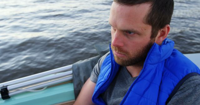Man relaxing on a rowboat, deep in thought, on a lake. He is wearing a blue vest and looking contemplative. Suitable for themes of reflection, nature, traveling, leisure activities, and emotional state. Ideal for articles, blogs, or campaigns centered on mental health, adventure, and outdoor activities.