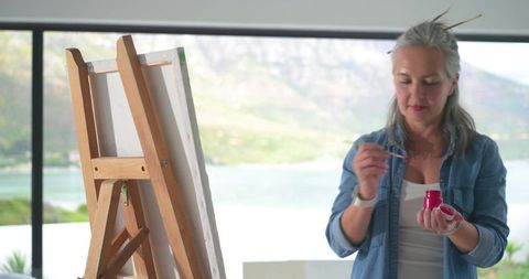 Mature woman mixing bright pink paint at waterfront art studio with easel and canvas