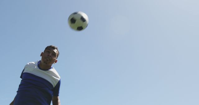 Male soccer player actively heading a soccer ball against clear blue sky background. Ideal for use in sports promotions, advertisements for athletic apparel or gear, or motivational sports materials showcasing focus and determination. Useful for illustrating articles on soccer techniques and athlete training.