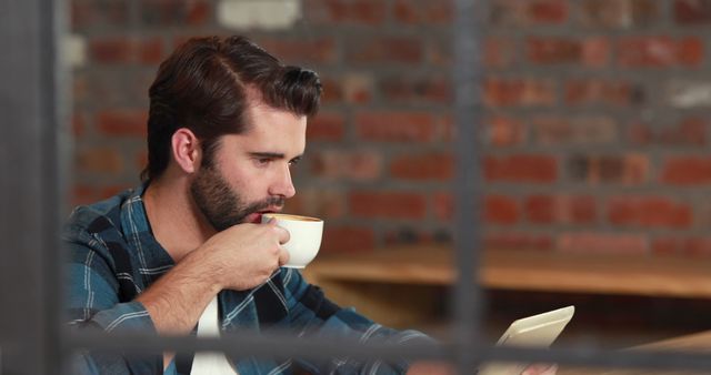 A young Caucasian man is focused on reading a tablet while sipping coffee in a cafe with a brick wall background, with copy space. His casual attire and the relaxed environment suggest a comfortable setting for work or leisure.