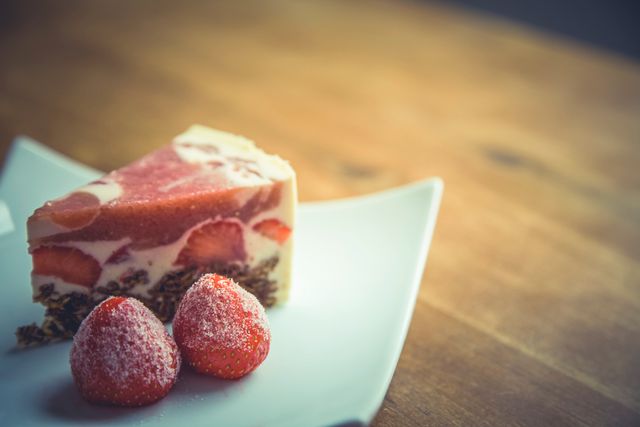 Strawberry cheesecake slice displayed on a white plate, complemented by two fresh strawberries, on a wooden table. Ideal for illustrating dessert recipes, gourmet food blogs, baking inspiration, food styling, and coffee shop menus.