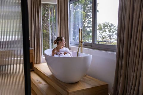 Woman relaxing in freestanding bathtub on wooden platform overlooking treetop view