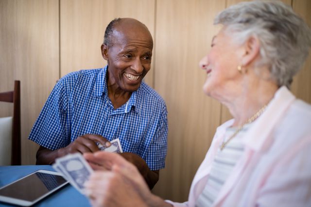 Senior male and female friends enjoying a game of cards at a table in a nursing home. They are smiling and engaging in a fun activity, showcasing companionship and happiness in a retirement setting. This image can be used for promoting elderly care services, retirement homes, and activities for seniors.