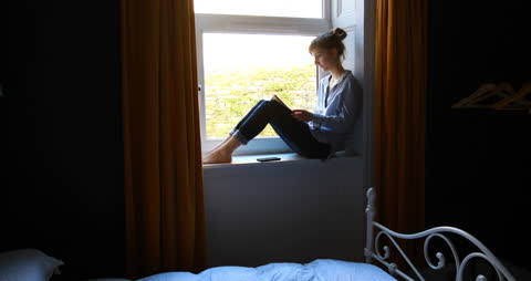 Woman Sitting on Window Sill Reading and Using Phone