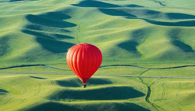 Vibrant red hot air balloon floating over rolling green farmland casting soft shadows