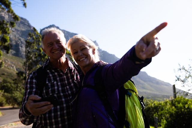Senior couple enjoying a hike in a scenic mountainous area. The woman is pointing at something while the man holds a smartphone. Ideal for use in advertisements promoting active lifestyles, retirement activities, travel, and nature exploration.