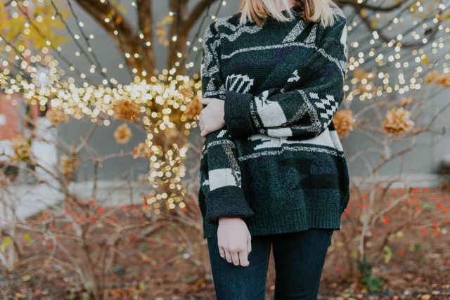 Young woman wearing a cozy sweater standing outside with holiday lights in the background. Ideal for use in holiday-themed content, fashion blogs, advertisements for winter apparel, or social media posts invloving seasonal activities and style.