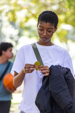 African american woman examining leaf in sunlit park, wearing glasses and holding jacket