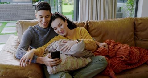 Diverse couple relaxing on cozy sofa browsing smartphone
