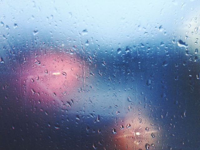 Raindrops splattered across a window pane obscure the view of blurred traffic lights in the background, evoking a moody and contemplative atmosphere. Suitable for use in projects or articles about weather, urban life, emotional themes, solitude, or rainy days.