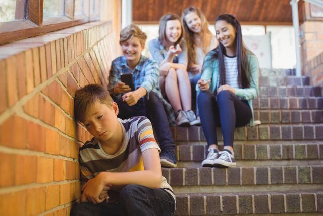 Image depicts a sad boy sitting alone on a staircase while a group of school friends are bullying him. This image can be used to illustrate topics related to bullying, peer pressure, emotional distress, and social issues among children and adolescents. It is suitable for educational materials, mental health awareness campaigns, and articles discussing the impact of bullying in schools.