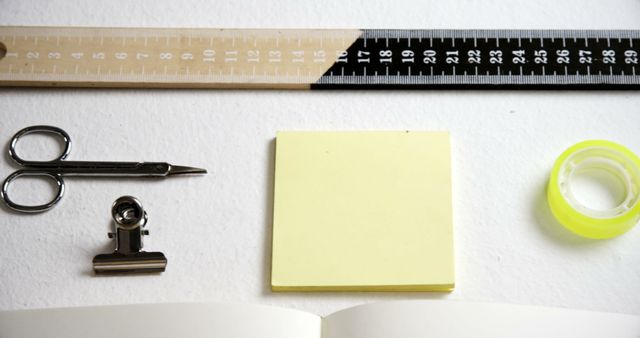 Top view of an organized arrangement of office supplies including a ruler, scissors, sticky notes, tape, and a clip. Use in business, school, organizational, or productivity-themed contexts.