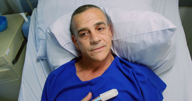 Man lying in hospital bed, wearing blue hospital gown, holding medical device on chest. Ideal for content related to healthcare, hospital, patient recovery, nursing, and medical services.