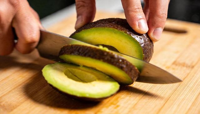 Slicing ripe avocado on wooden cutting board with kitchen knife close-up of hands