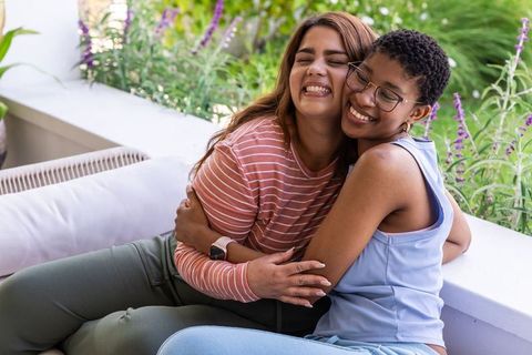 Female friends hugging and smiling on sunlit porch bench with cushions and greenery