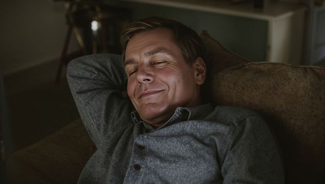 Middle-aged man relaxing on couch at home in cozy living room with warm low lighting