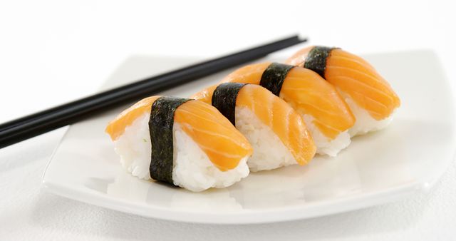 Three salmon nigiri pieces arranged neatly on a white plate with a pair of black chopsticks in the background. Ideal for promoting Japanese cuisine, sushi restaurants, culinary publications, and food blogs.