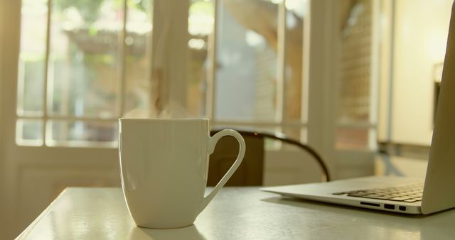 Warm cup of coffee sits on a table next to an open laptop in a sunlit room with large windows. Ideal for illustrating concepts of morning routines, home workspaces, relaxation, or cozy atmospheres.