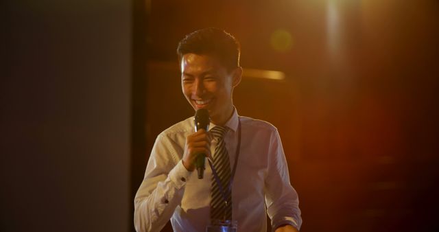 Young businessman in a white shirt and striped tie holding a microphone and smiling while delivering a presentation or speech in a dimly lit room with a warm light flare. Suitable for themes related to corporate events, public speaking, business presentations, and leadership.