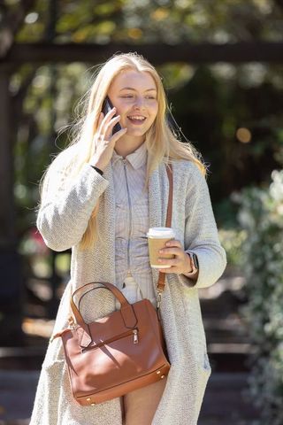 Young woman walking and talking on phone while holding takeaway coffee and tan handbag