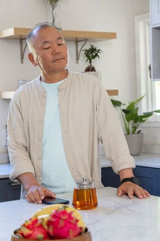 Mature middle-aged asian man enjoying tea in sunlit modern minimalist kitchen by marble island