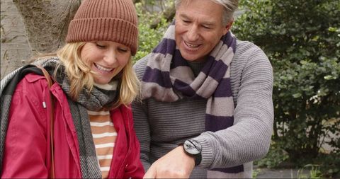 Senior couple sharing smartphone and laughing on park bench wearing warm fall clothing