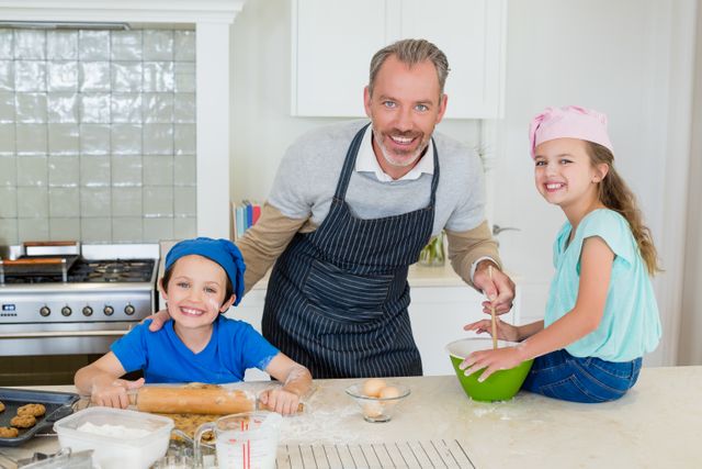 Father and children enjoying baking together in a modern kitchen. The father is wearing an apron and smiling while mixing ingredients in a bowl. The children are also smiling, with one rolling dough and the other sitting on the counter. Ideal for use in advertisements, family-oriented content, cooking blogs, and lifestyle articles emphasizing family bonding and home activities.