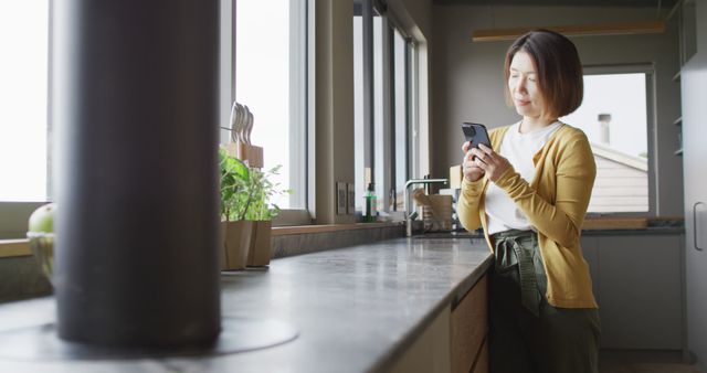 Asian woman standing in modern, bright kitchen during daytime, using smartphone while leaning on counter. This image is ideal for promoting home and lifestyle products, technology, communication services, or depicting leisure and relaxation in a domestic environment.