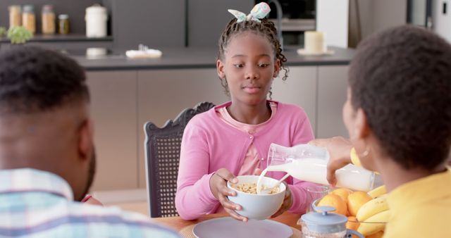 Young girl in pink cardigan eating breakfast with her family in a modern kitchen. She is holding a bowl of cereal while someone pours milk into it. Ideal for depicting family life, morning routines, domestic happiness, and healthy eating. Suitable for ads or articles on family bonding, nutritional tips, and home life.