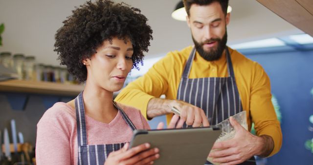Image of happy diverse couple in aprons using tablet and baking in kitchen at home. Happiness, inclusivity, free time, togetherness and domestic life.