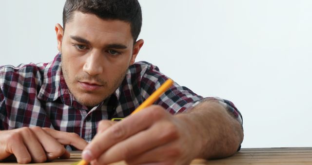 Young man is concentrating while measuring with a pencil on a wooden surface, wearing plaid shirt. Useful for representations of precision work, craftsmanship, DIY activities, woodworking, and dedication.