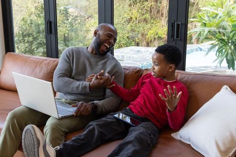 African american father and son laughing on couch bonding with laptop and tablet