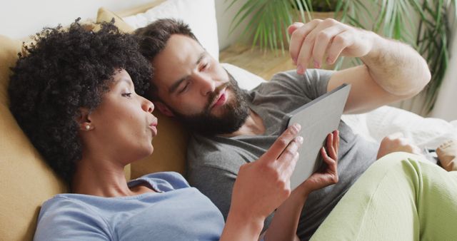 Image of happy diverse couple relaxing at home, lying on bed using tablet. Happiness, communication, inclusivity, free time, togetherness and domestic life.