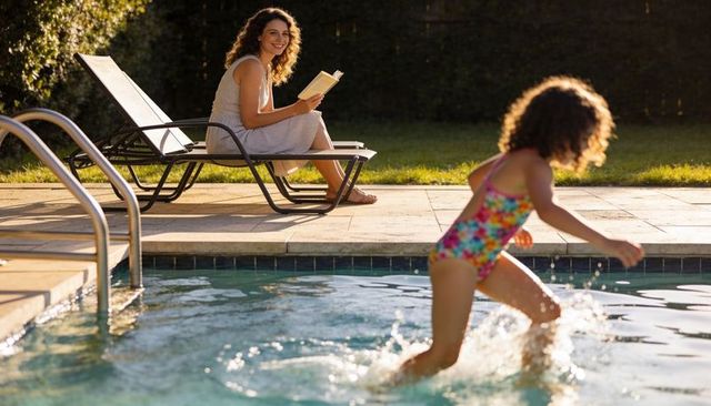 Mother reading on poolside lounger while daughter splashing in sunlit backyard pool at golden hour