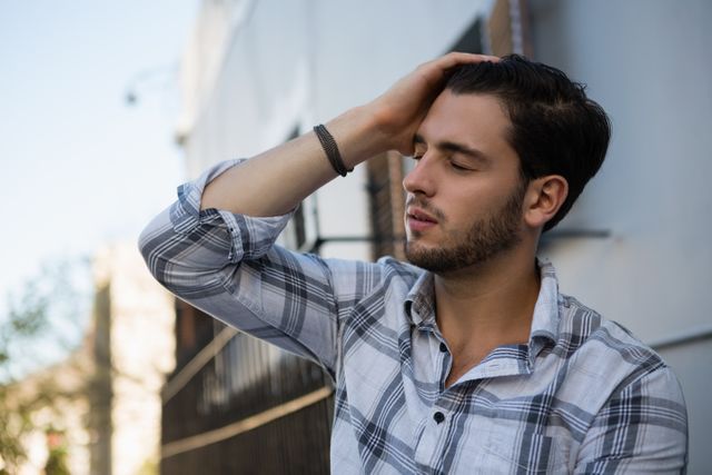 Young man in casual plaid shirt leaning against wall with eyes closed and hand on head, appearing stressed or deep in thought. Suitable for illustrating concepts of stress, contemplation, emotional struggle, or urban lifestyle.