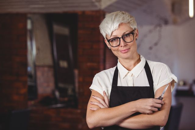 Female hairdresser standing confidently with arms crossed in a modern salon, wearing an apron and glasses. Ideal for use in articles or advertisements related to beauty industry, hairdressing services, professional hairstylists, and salon promotions.