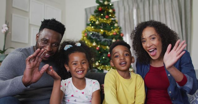 African American family is at home, joyfully waving at camera during Christmas celebrations. Parents and children are smiling, looking happy together around Christmas tree adorned with decorations, capturing joyful holiday moments. Ideal for family holiday greetings, advertisements featuring cultural traditions, visuals representing festive home environments, or use in content related to family bonding during the holidays.