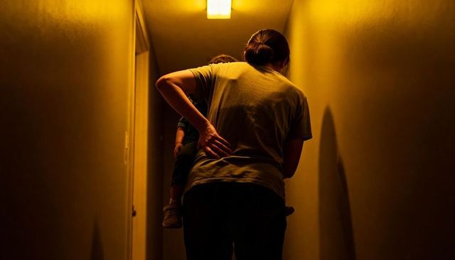 Parent carrying tired child in narrow dimly lit hallway backlit by warm ceiling light