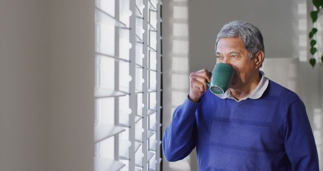 Image of senior biracial man standing looking out of window and drinking coffee. Retirement, healthcare, inclusivity and senior lifestyle concept.
