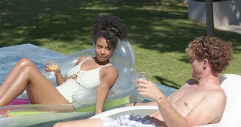 Relaxing couple lounging on pool floats sipping drinks in sunlit backyard