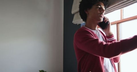 African american man talking on phone while adjusting window blind in minimalist interior