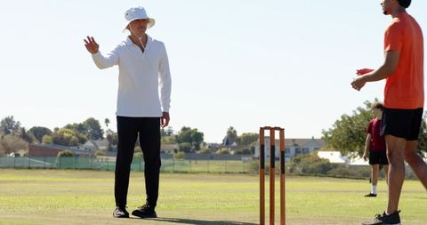 Cricket umpire signaling out wearing bucket hat and white shirt near stumps on pitch
