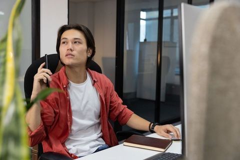 Young asian professional talking on smartphone at modern office desk with notebook and monitor